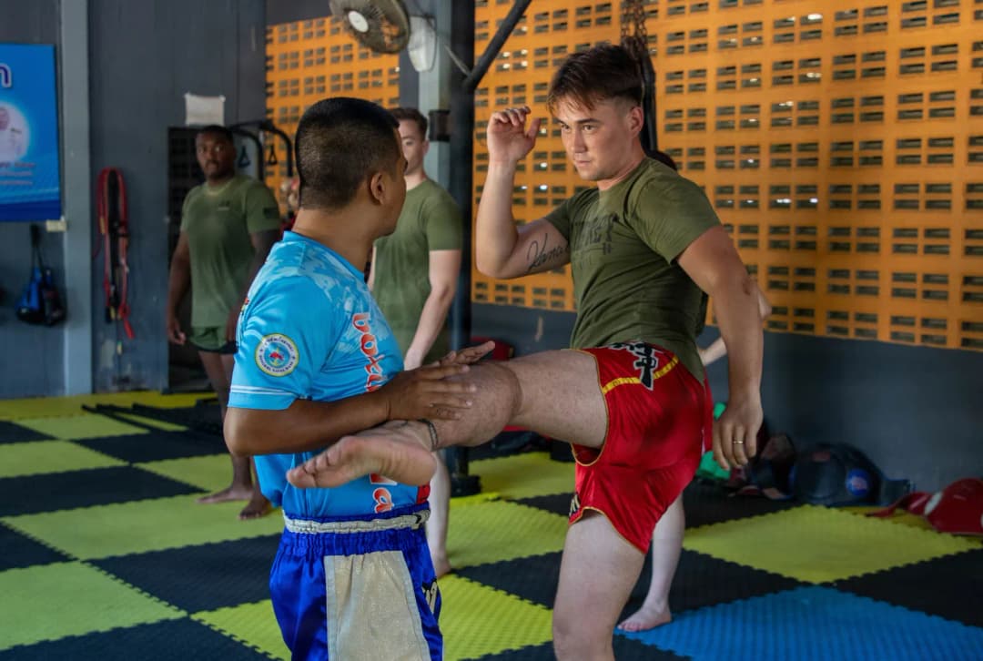 Muay Thai trainer in blue shorts guiding a student through a kick on padded gym mats